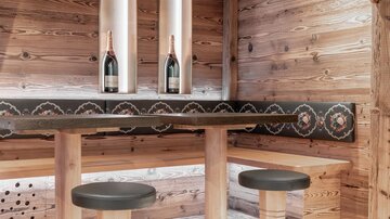 A cozy wooden bar with two stools and a thick tabletop. In the background, two bottles of champagne are placed in elegant holders.