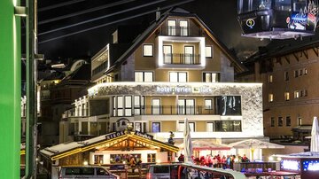 A modern hotel with a illuminated facade in an Alpine town at night. Next to the hotel are a souvenir stand and people enjoying themselves.