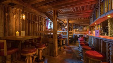 A cozy bar with wooden paneling and warm lighting. The decor consists of tables and chairs with red upholstery.