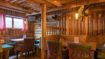 A cozy restaurant with wooden walls and rustic design.  
The seating is adorned with colorful fabrics and there are windows with a view outside.