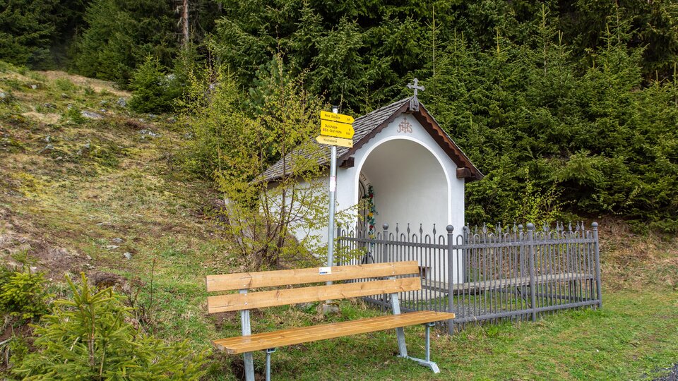 A small chapel stands in nature, surrounded by green trees. Next to the chapel is a wooden bench for sitting and resting. | © Bernhard Gruber – Photography