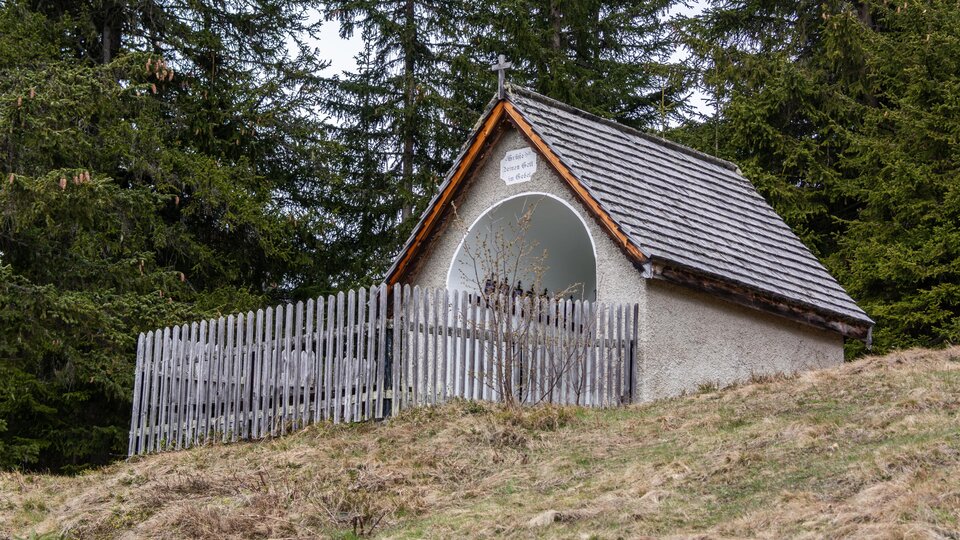 A small hut with a wooden fence in a wooded area. The sky is cloudy, and the ground is covered with grass and leaves. | © Bernhard Gruber – Photography