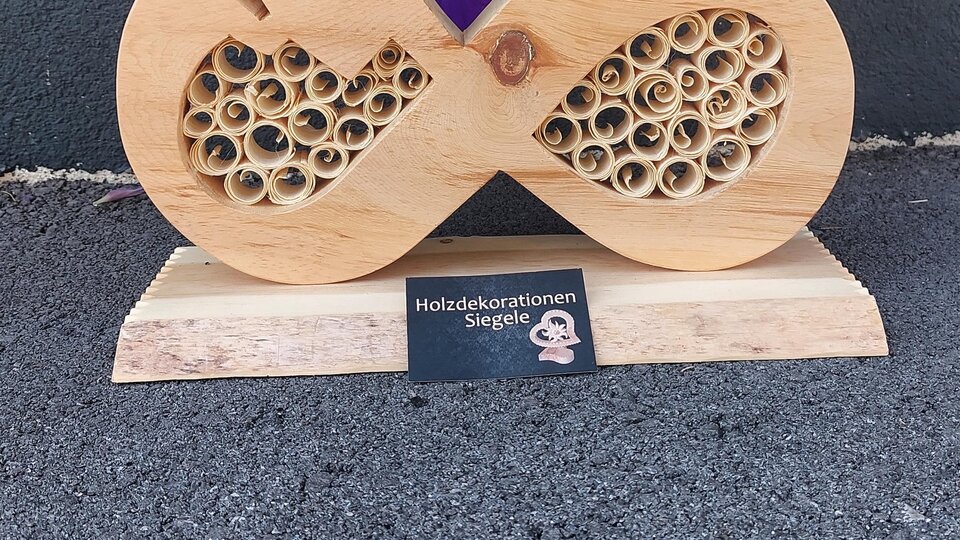 A decorative wooden sculpture in the shape of an infinity symbol with heart motifs and a spiral pattern. In the background, there are purple flowers.