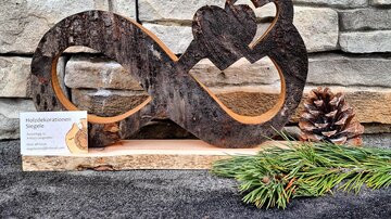 A wooden sculpture in the shape of an infinity loop with a heart. Next to the sculpture lie a pine cone and some pine needles.