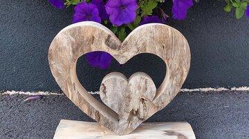 A wooden sculpture in the shape of a heart with a smaller heart in the center. In the background, purple petunias are blooming.