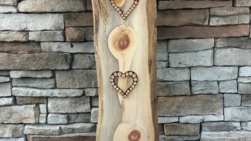 A decorative wooden block with two heart-shaped cutouts. In the background, colorful flowers and a stone wall are visible.