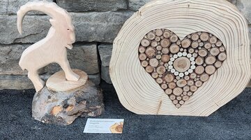 A handcrafted wooden sculpture set. It features a goat figure and a heart made of wooden discs on a stone wall.