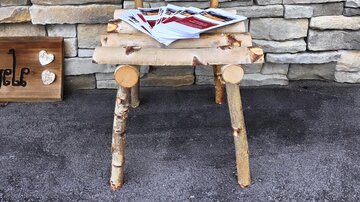A rustic chair made of wooden logs stands in front of a stone wall. There are some magazines on the chair.
