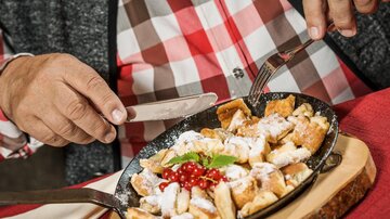 A plate of freshly prepared Kaiserschmarrn, garnished with powdered sugar and red currants. A person is cutting the sweet dish with a knife and fork.