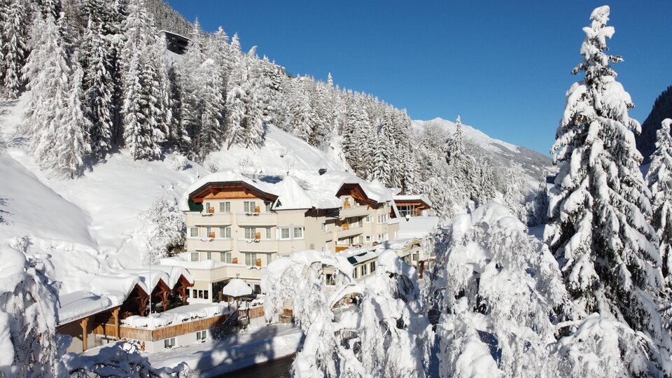 A picturesque hotel in a snow-covered winter landscape. Tall pine trees and a clear blue sky surround the scene.