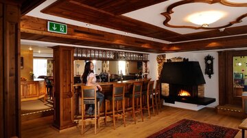 A cozy bar with wooden paneling and an open fireplace. In the background, a woman is standing at the bar and there are several seating areas.