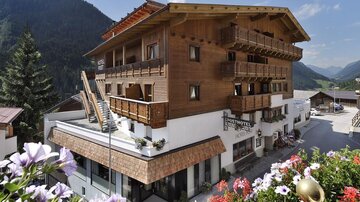 A charming wooden building with several balconies and blooming flowers at the entrance. In the background, picturesque mountains and a clear sky can be seen.
