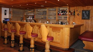 A cozy bar with wooden paneling and high chairs. Behind the bar are various alcoholic beverages and glasses.