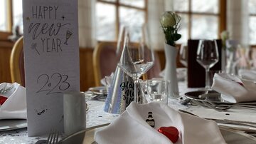 A festively set table for New Year's Eve with elegant plates, glasses, and decorated napkins. In the background stands a table with the inscription "Happy New Year 2023".