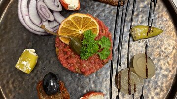 An artistically arranged beef steak tartare with onions, lemon slice, and various side dishes. The plate also features black olives and a sophisticated sauce.