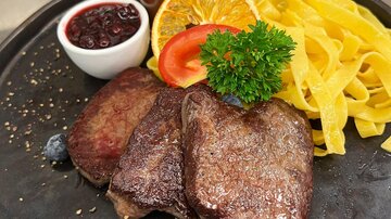 An elegantly arranged plate with juicy steak, fresh egg noodles, and sides of orange and tomato. Accompanied by a small bowl of cranberries and a sauce.