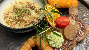 An artfully arranged plate with meat, a sauce, and various side dishes. Accompanied by filled croquettes, fresh vegetables, and garnish.