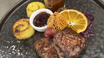 An appealing plate with grilled meat, decorated with an orange slice and a crunchy dessert. Alongside a portion of sauce and sides of polenta and served fruit.