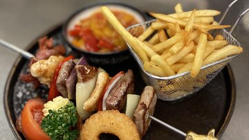 A plate with grilled meat, vegetables, a breaded onion, and potato wedges. It comes with French fries and a spicy sauce.