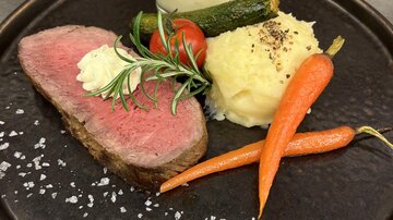 A delicious dish with pink roasted beef, sides of mashed potatoes, carrots, and zucchini. A glass with a creamy sauce completes the meal.