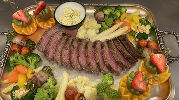 An artistically arranged meat platter with perfectly cooked beef steak, surrounded by colorful vegetables and fresh fruits. In the center, there is a bowl of sauce.