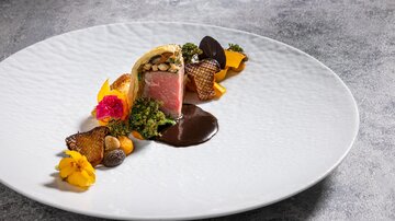 An elegant dish on a white plate, consisting of meat, side dishes, and decorative edible flowers. The arrangement is artistic and appealing.