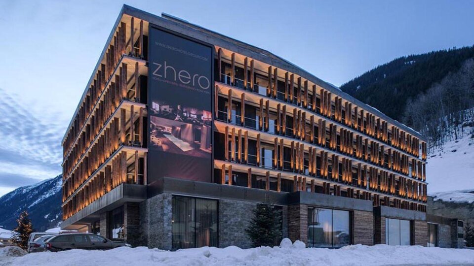 A modern hotel building with wooden cladding and large windows. Surrounded by snow-covered mountains and a clear sky.