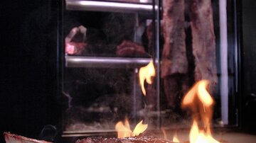 A juicy steak sizzles on a grill, while flames flicker around. In the background, large pieces of meat are visible.