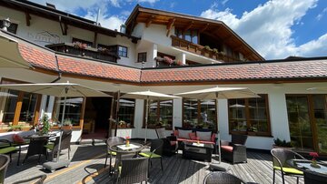 A cozy outdoor terrace of a restaurant with sun umbrellas and stylish furniture. In the background, there is a modern building with wooden decorations and blooming windows.
