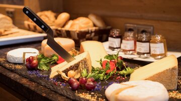 Ein ansprechendes Käsebrett mit verschiedenen Käsesorten, Trauben und frischem Grün. Im Hintergrund sind Brötchen und Marmeladegläser zu sehen.