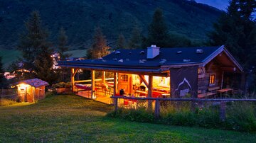Ein gemütliches Chalet in den Bergen bei Dämmerung. Der beleuchtete Innenraum und die umliegende Natur schaffen eine einladende Atmosphäre.