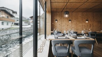 A modern restaurant with wooden paneled walls and large windows. The tables are elegantly set and offer a view of the surroundings.