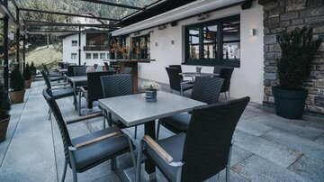 A stylish outdoor area of a restaurant with tables and chairs. In the background, mountains and a peaceful setting can be seen.