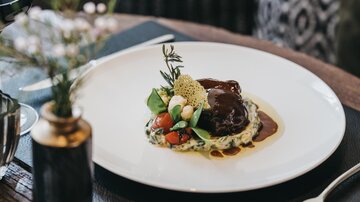 An elegantly arranged dish on a white plate with a dark sauce. Accompanied by fresh vegetables and herbs.