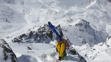 A skier in a yellow jacket is standing in the snow and looking down the sloped descent. In the background, snow-covered mountains can be seen.