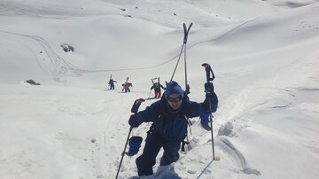 A ski mountaineer climbs a snow-covered slope with ski poles. In the background, there are more people who are also attempting the ascent.