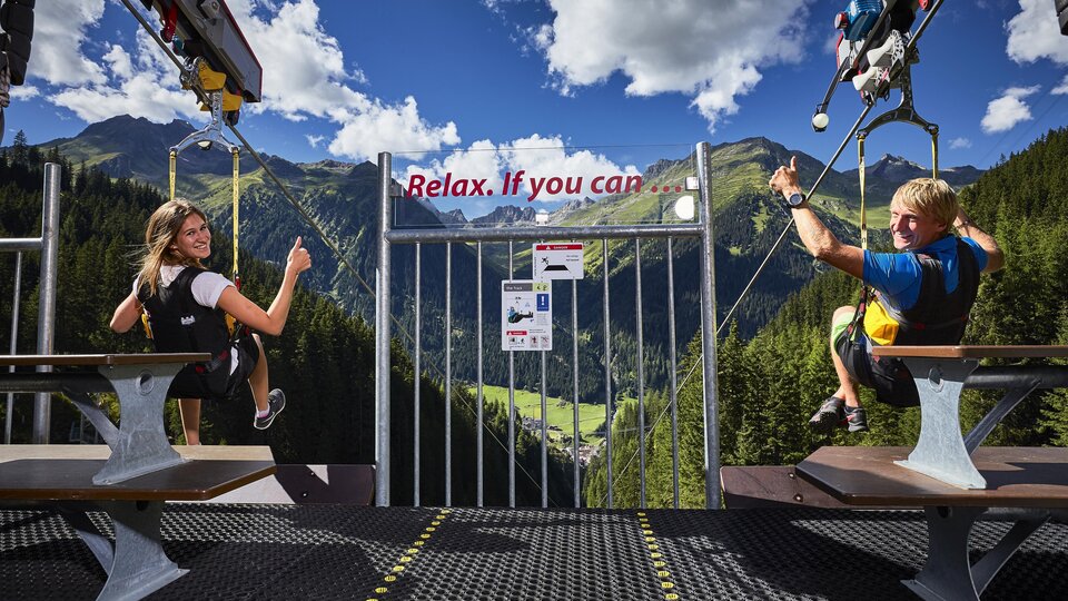 Two people are sitting on a platform overlooking a picturesque mountain landscape. A sign encourages relaxation.