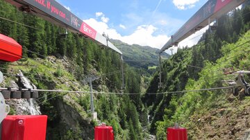 A cable car with a view of a green ravine. The sky is blue with some clouds.