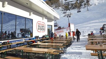 A modern mountain hut café with outdoor seating, surrounded by snow-covered mountains. Skiers and snowboarders enjoy the day in a winter paradise.