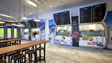 A modern café with light wood chairs and large windows. On the wall, there are advertising boards and an appealing counter with colorful drinks and snacks.