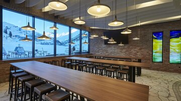 A modern restaurant with large windows overlooking the snow-covered mountains. The space is brightly lit and features long wooden tables and chairs.