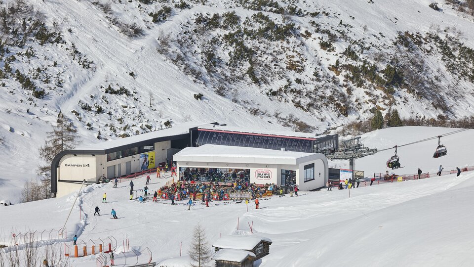 A bustling ski resort in the mountains with many skiers. In the background, snow-covered slopes and a cable car can be seen.