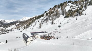 A picturesque snow landscape with a ski lift and a mountain lodge. In the background, there are snow-covered mountains and a blue sky.