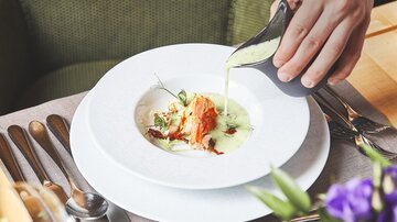 An elegant dish is poured into a white plate. In the background, cutlery and a floral arrangement can be seen.