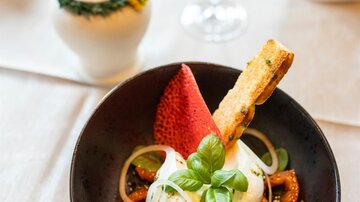 A stylish dish on a black plate, presented with fruits and fresh herbs. Next to the plate, there is a glass and a decorative lamp.