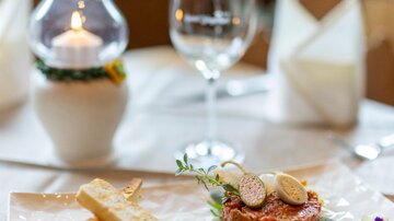 An elegant dish on a white plate, served with fresh sides. In the background, there is a glass of wine and a candlelight.