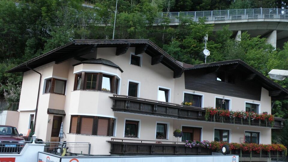 A charming building with several balconies and blooming windows. In the background, trees and a street bridge are visible.