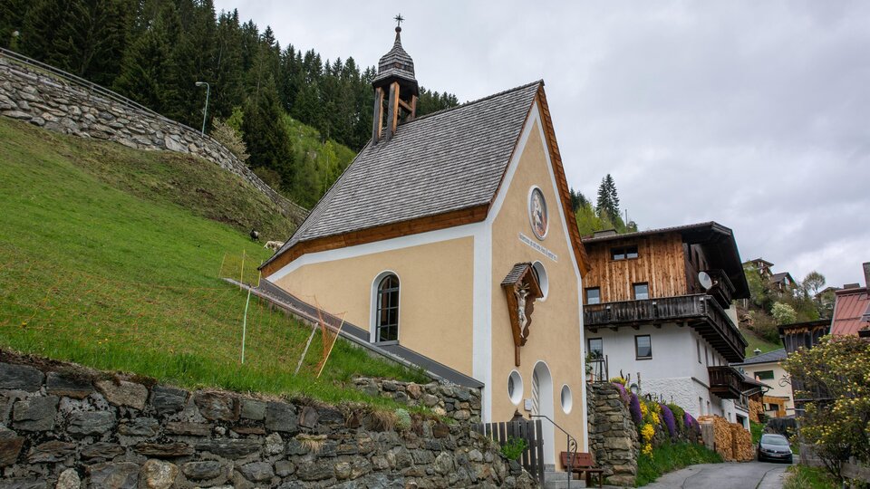 A small, orange chapel stands on the slope, surrounded by greenery. In the background, typical alpine wooden houses can be seen. | © Bernhard Gruber – Photography