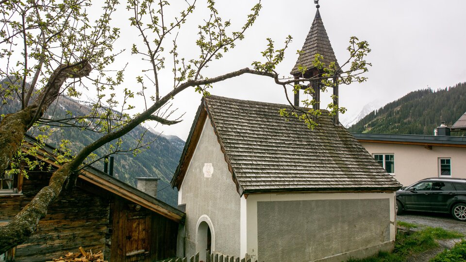 A small chapel with a pointed roof and a bell tower is surrounded by trees and mountains. In the foreground, there is a wooden fence. | © Bernhard Gruber – Photography
