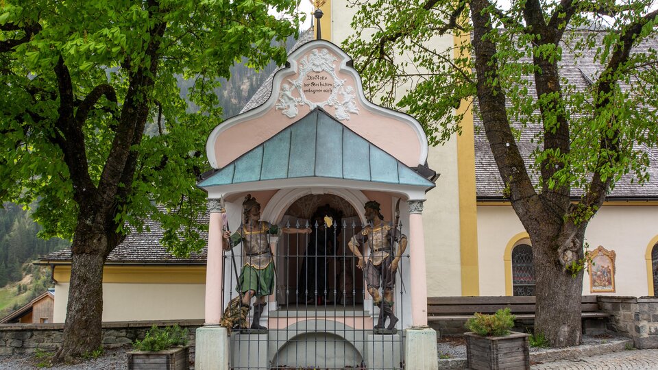 A small chapel with colorful figures and a green roof. In the background, there are large trees and a building with a yellow facade. | © Bernhard Gruber – Photography